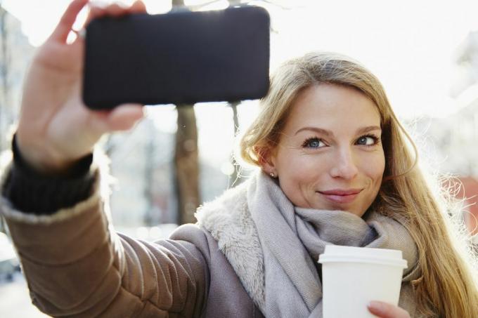 čedna ženska, ki pozimi hodi po mestnih ulicah, posname selfi in drži kavo za s seboj, osvetljeno od zadaj