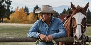 kevin costner v Yellowstonu poleg konja, ki se naslanja na ograjo z vrvjo v rokah, oblečen v obledelo modro denim srajco in bež kavbojski klobuk