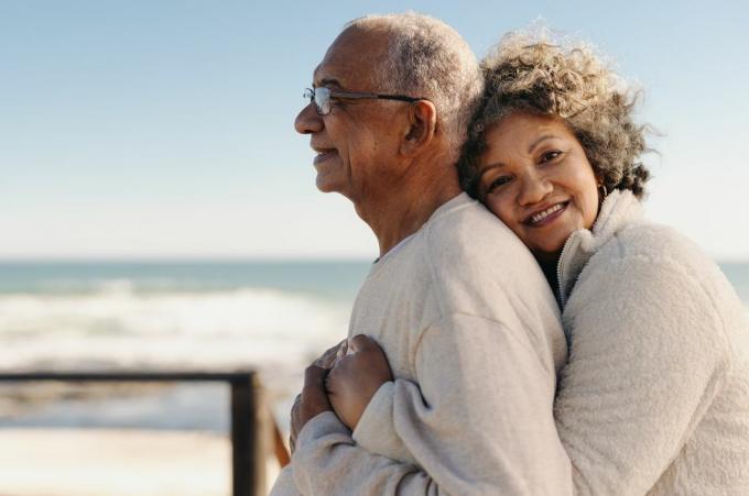 romantična starejša ženska se smehlja v kamero, medtem ko objema svojega moža ob oceanu ljubeč starejši par, ki po upokojitvi uživa skupaj preživeti nekaj kakovostnega časa